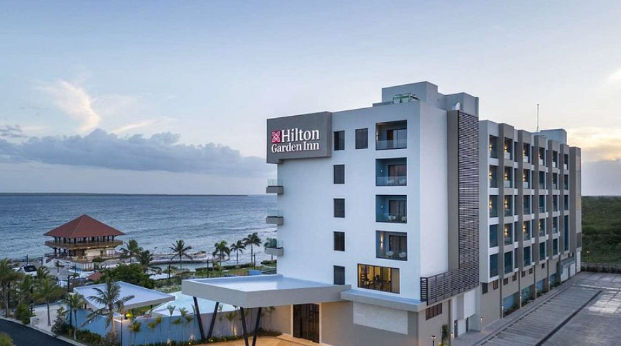 Exterior photograph of the modern, multi-story Hilton Garden Inn hotel building by the sea at sunset. The building has light-colored walls and numerous balconies with sea views. The entrance has a grey awning, and there are small buildings, palm trees, an