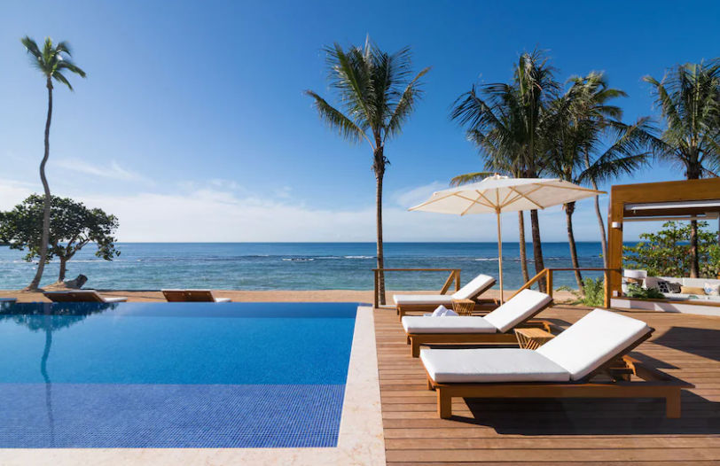 Infinity pool facing the ocean with wooden lounge chairs, a white umbrella, and palm trees under a clear blue sky