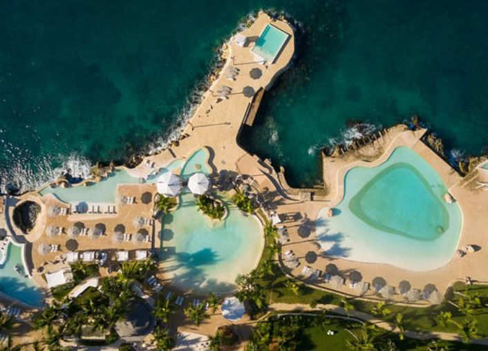 Una vista aérea de dron de un lujoso resort costero situado en una costa rocosa. Muestra múltiples piscinas de agua azul claro con formas orgánicas y bordes de piedra, rodeadas de numerosas tumbonas, sombrillas de playa y palmeras. Un espigón o muelle de 