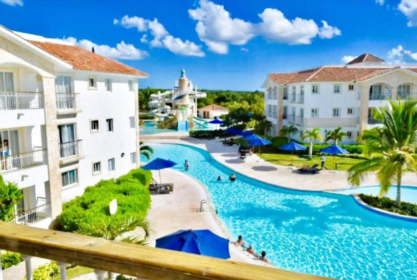 Aerial view of the swimming pool at Cadaques Caribe Resort with guests, surrounded by white villas and palm trees under a clear sky