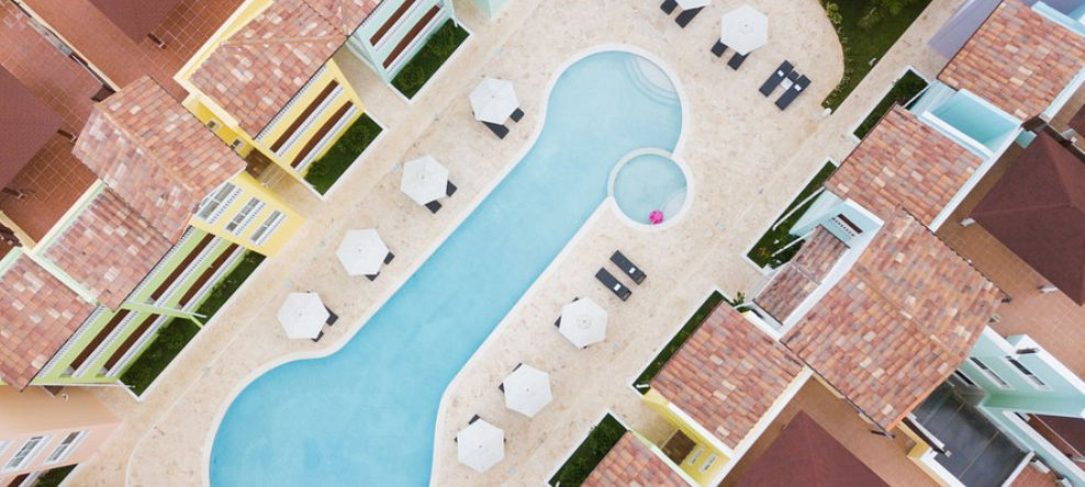 Vista aérea de un complejo hotelero que muestra una piscina alargada de color azul claro, rodeada por una terraza con sombrillas blancas, tumbonas y edificios de colores pastel con techos de tejas de terracota.