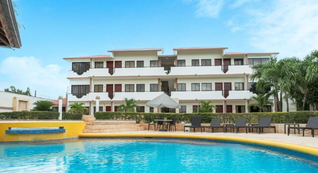 A large, curved swimming pool with clear blue water and a yellow edge in the foreground. Next to the pool, black chaise lounge chairs and a patio table with a closed grey umbrella are arranged. Steps lead up to a low green hedge. Beyond the hedge is a thr