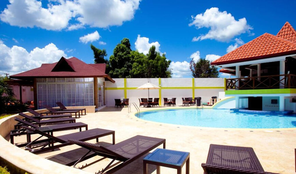 Tumbonas se alinean en el primer plano de una gran piscina de complejo turístico bajo un cielo parcialmente nublado, con áreas de comedor cubiertas y un bar en la piscina en el fondo