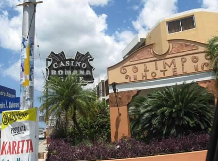 Exterior facade of Hotel Olimpo in La Romana featuring the Casino Romana sign in the background under a partly cloudy sky.