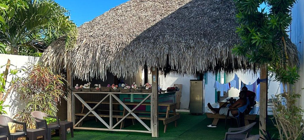 Área recreativa al aire libre del Hostal Johnnier Macao con una estructura de techo de paja, sillas de plástico dispuestas en el césped artificial y la fachada colorida del edificio del hotel al lado.