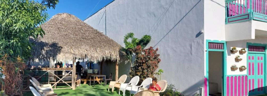 Outdoor recreational area of Hostal Johnnier Macao with a thatched roof structure, plastic chairs arranged on artificial turf, and the colorful facade of the hotel building next to it."
