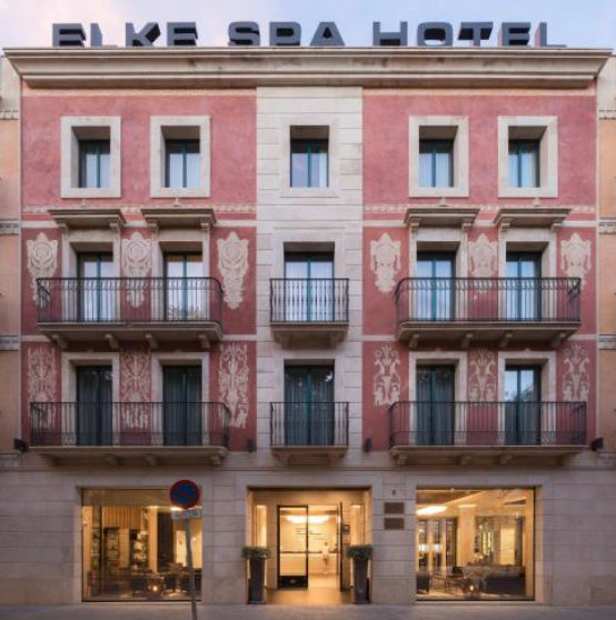 Facade of a pink and cream stone building with ELKE SPA HOTEL sign on top. It features black iron balconies on three floors above large ground-level windows.