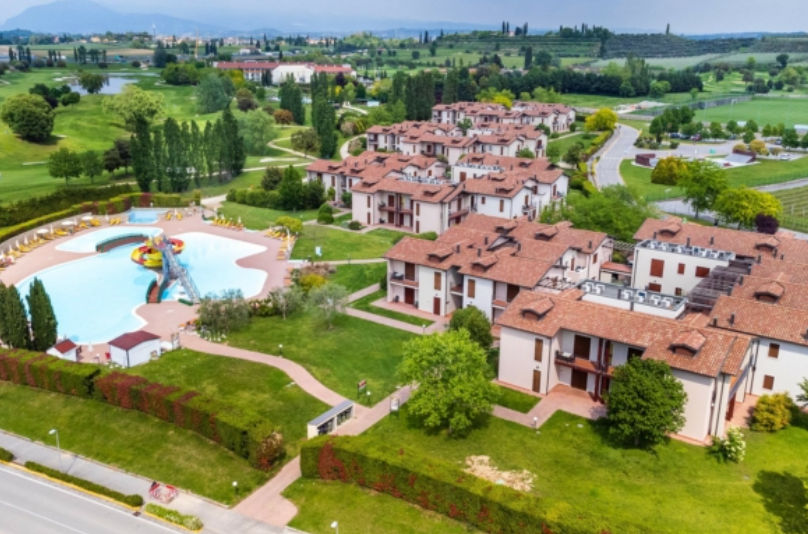 Aerial view of a holiday resort featuring red-roofed buildings, extensive green lawns, and a large swimming pool with a water slide surrounded by lounge chairs.