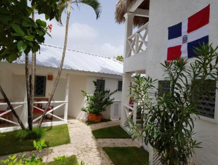 Patio interior de un hotel con casas blancas, palmeras, un camino de piedra y la bandera de la República Dominicana colgada en la pared.