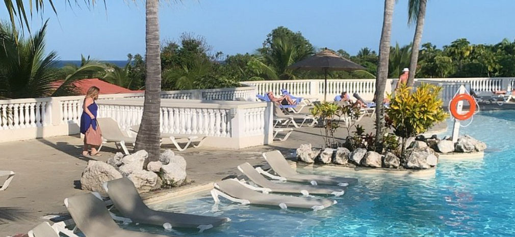 Área de la piscina al aire libre en un complejo turístico tropical con palmeras, sillones reclinables, sombrillas, un bar en la piscina y edificios de hotel de varios pisos bajo un cielo azul despejado