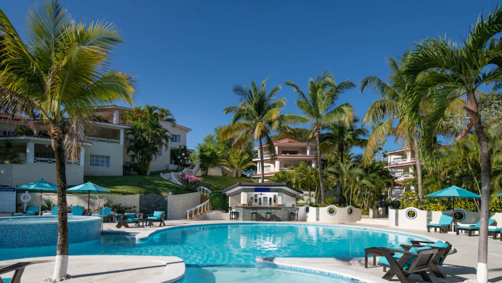 Área de la piscina al aire libre en un complejo turístico tropical con palmeras, sillones reclinables, sombrillas, un bar en la piscina y edificios de hotel de varios pisos bajo un cielo azul despejado.