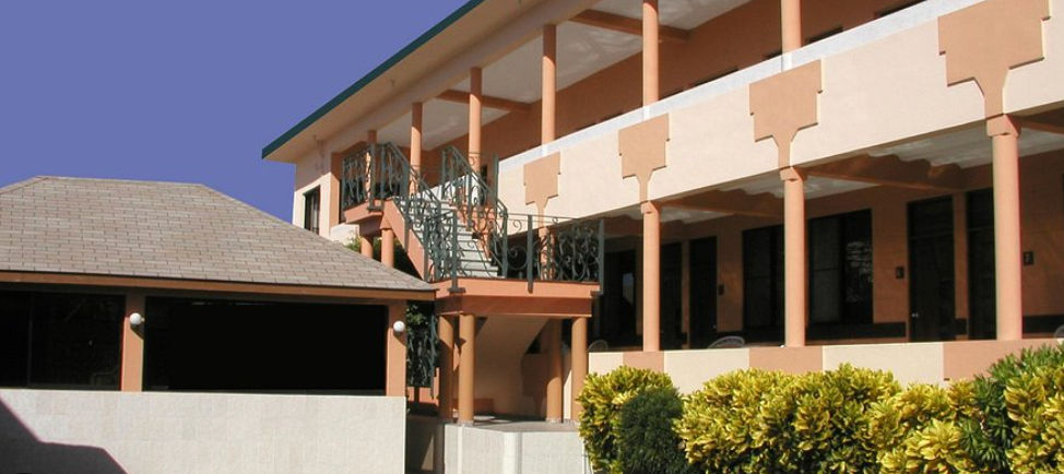 Two-story exterior facade of Hotel Garant & Suites featuring open hallways, peach-colored columns, ornate metal stairs, and green bushes in the foreground under a clear blue sky.