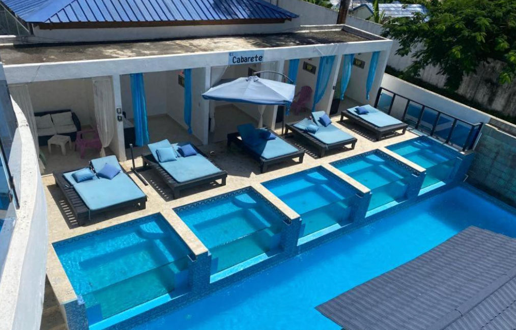 Un edificio de hotel blanco de tres pisos con balcones que tienen barandillas de vidrio teñido azul y muebles de exterior, con una choza de paja y árboles de palma a la izquierda, bajo un cielo azul claro