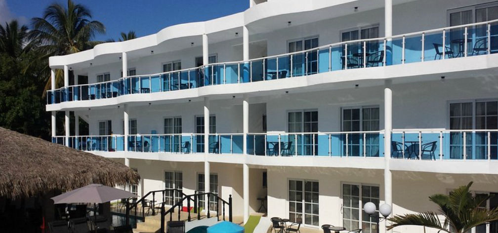 Un edificio de hotel blanco de tres pisos con balcones que tienen barandillas de vidrio teñido azul y muebles de exterior, con una choza de paja y árboles de palma a la izquierda, bajo un cielo azul claro.