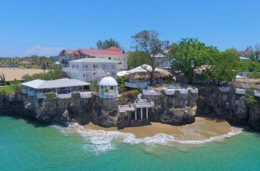 depicts a coastal hotel in Negril, Jamaica, it could have an alt text such as "Coastal resort in Negril, Jamaica, featuring white buildings with red roofs, a private beach on turquoise Caribbean water, and a white gazebo. It also includes white stairs lea