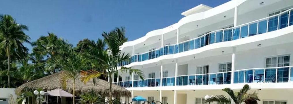 Edificio de hotel blanco moderno con balcones de vidrio azul junto a la piscina con techo de paja en el Caribe
