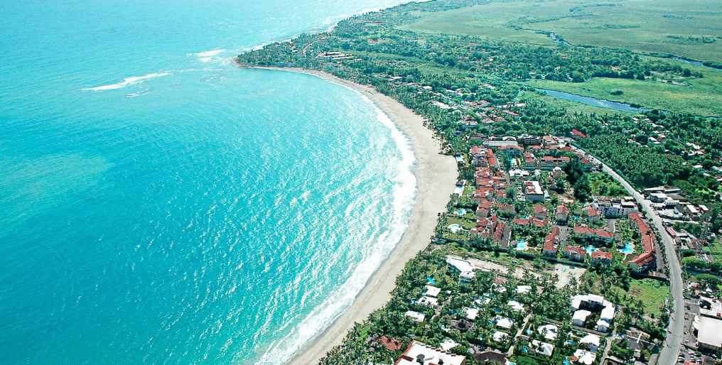 Una vista aérea de ángulo alto de una ciudad costera tropical, con aguas oceánicas turquesas, olas blancas y una playa de arena a la izquierda, que transiciona a edificios con techos rojos, vegetación verde, palmeras, una carretera y árboles densos de col