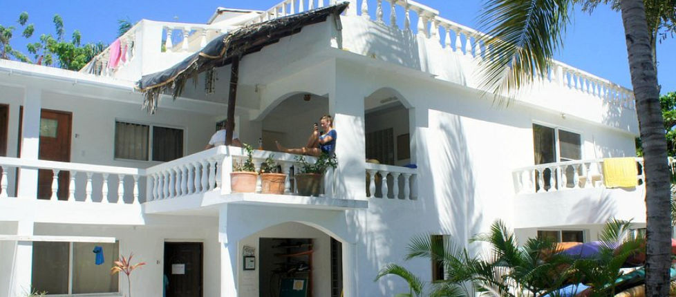 White exterior facade of The Hummingbird Cabarete featuring balconies, tropical plants, and palm trees under a clear blue sky"
