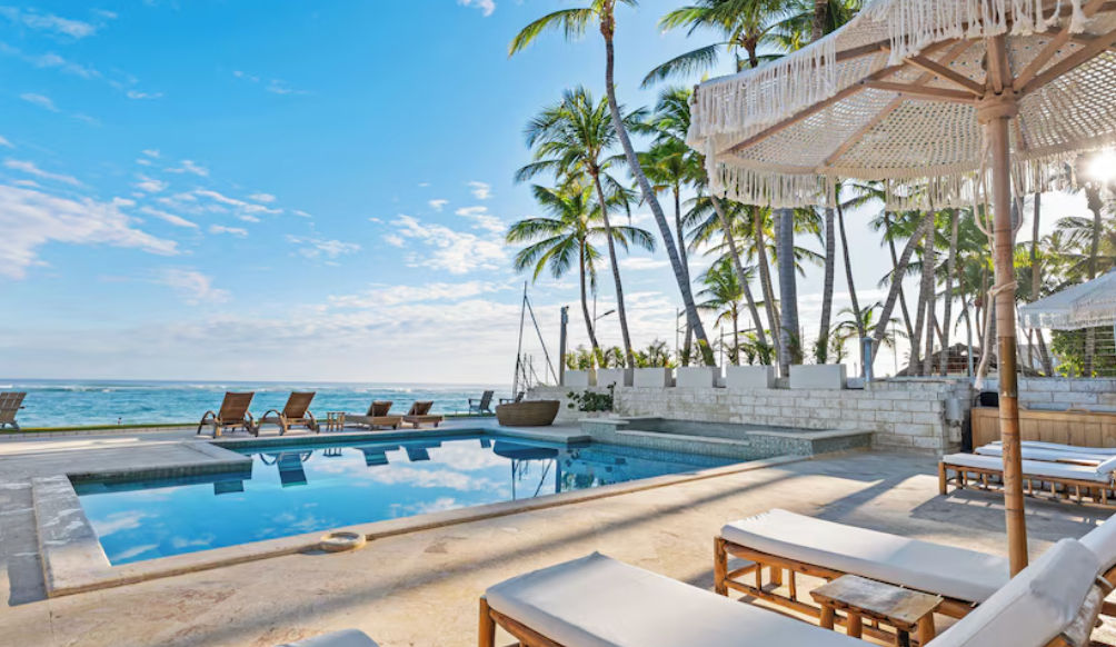 Exterior night shot of Watermark Luxury Oceanfront Residences at dusk, showing illuminated balconies, a swimming pool, and lounge chairs. Palm trees frame the building against a twilight sky.