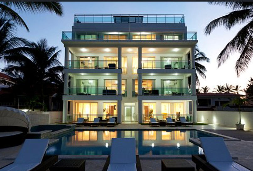 Exterior night shot of Watermark Luxury Oceanfront Residences at dusk, showing illuminated balconies, a swimming pool, and lounge chairs. Palm trees frame the building against a twilight sky.