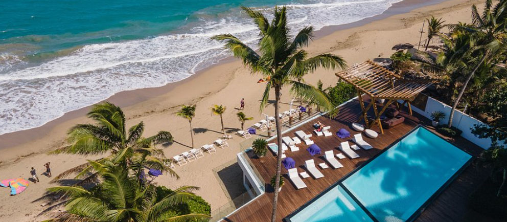 Aerial view of the infinity pool and wooden deck at Ultravioleta Boutique Residences facing Cabarete beach with palm trees.