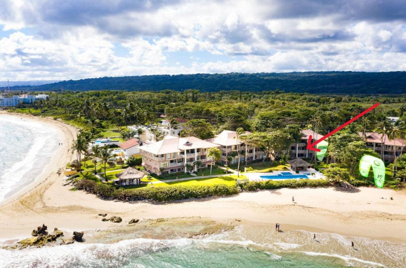 Vista aérea desde gran altura de un complejo residencial y resort de playa de varios pisos en una ubicación costera tropical. Grandes edificios con techos de terracota están rodeados de palmeras, césped verde y dos piscinas. Una amplia playa de arena con 