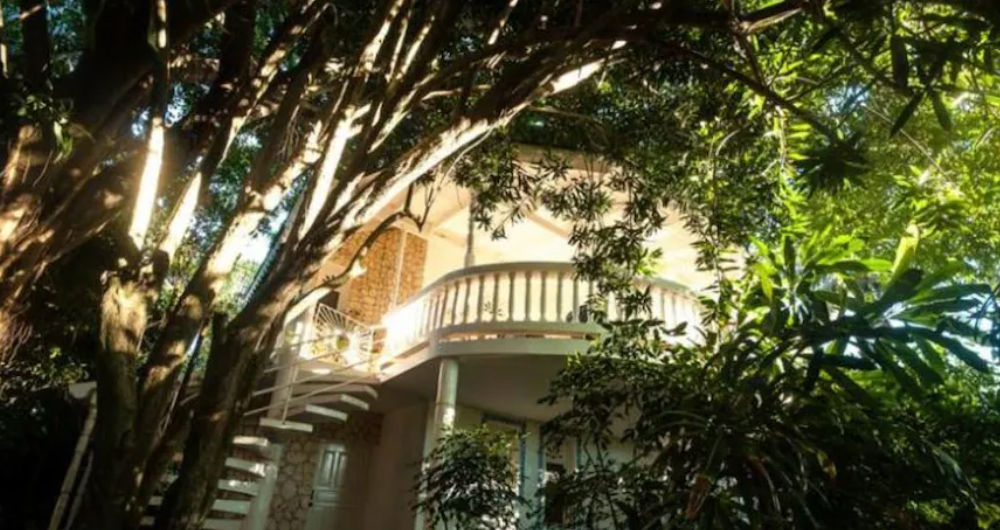 Two-story tropical cabin surrounded by dense trees with a white curved balcony and outdoor spiral staircase