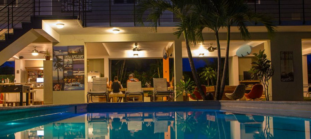 Escena nocturna en un campamento de surf tropical que muestra una piscina iluminada en primer plano con una sala común cubierta y un área de cocina, personas sentadas en una mesa, palmeras y un edificio de varios niveles con un balcón superior al fondo. U
