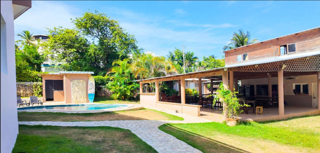 Un patio interior tropical con piscina, una gran área de comedor con techo de madera y palmeras exuberantes bajo un cielo despejado.
