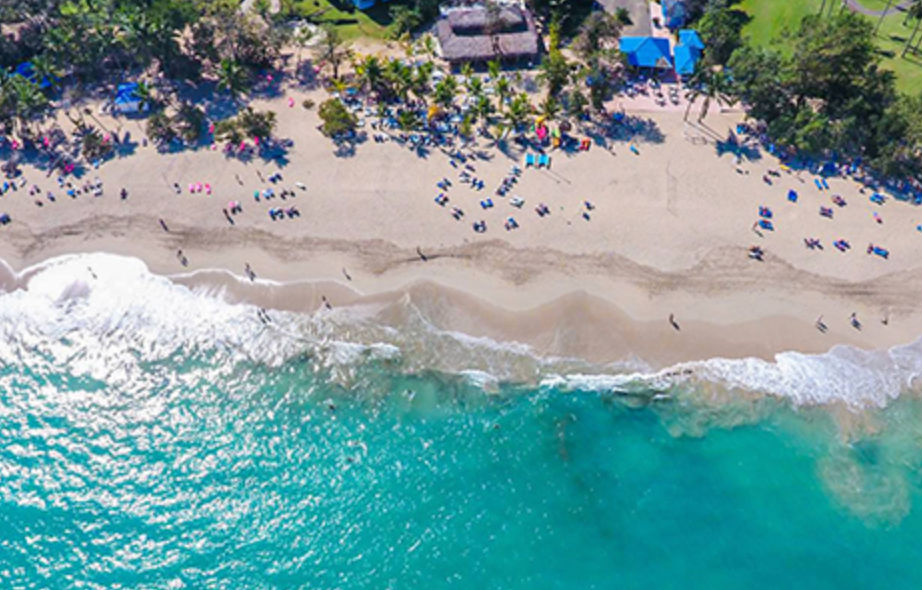 Vista aérea de un complejo turístico tropical en la costa con piscina turquesa, palmeras, chozas con techo de paja y zona de playa junto al mar