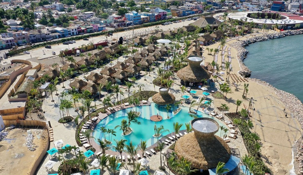 Aerial view of a tropical coastal resort featuring a turquoise swimming pool, palm trees, thatched-roof huts, and a beach area next to the ocean.