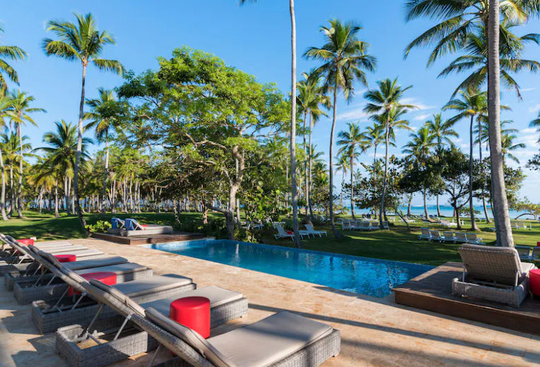 Piscina tropical rodeada de palmeras con tumbonas y una pérgola blanca en un resort de Punta Cana