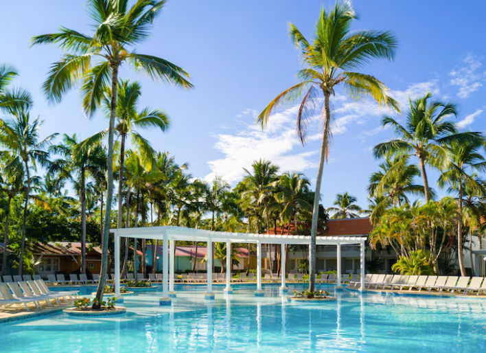 Piscina tropical rodeada de palmeras con tumbonas y una pérgola blanca en un resort de Punta Cana.