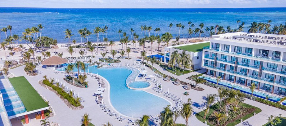 Vista aérea del Serenade Punta Cana Beach & Spa Resort destacando su amplia piscina central, instalaciones modernas, palmeras tropicales y la playa de aguas turquesas al fondo.