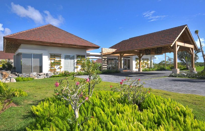 Main entrance and reception area of Los Establos Cap Cana featuring rustic wooden roofs, a paved driveway, and a tropical garden with green shrubs and pink flowers under a clear blue sky.