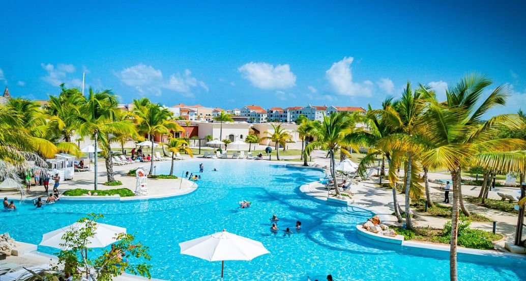 Mediterranean-style resort buildings in pastel blue and yellow at Ancora Punta Cana, featuring palm trees and a swimming pool in the foreground