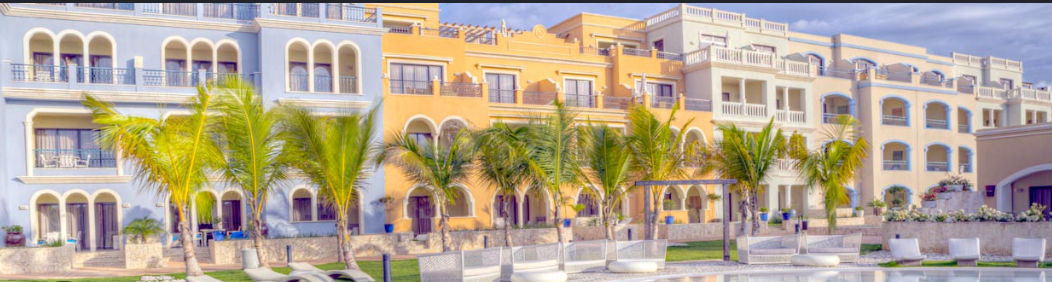 Mediterranean-style resort buildings in pastel blue and yellow at Ancora Punta Cana, featuring palm trees and a swimming pool in the foreground.