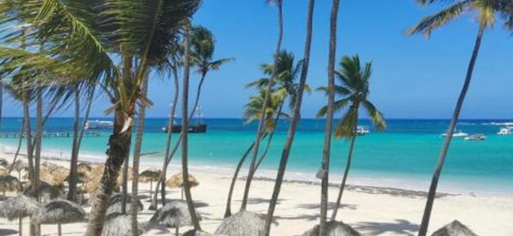 Playa tropical de arena blanca con palmeras, sombrillas de paja y agua cristalina color turquesa con barcos al fondo.