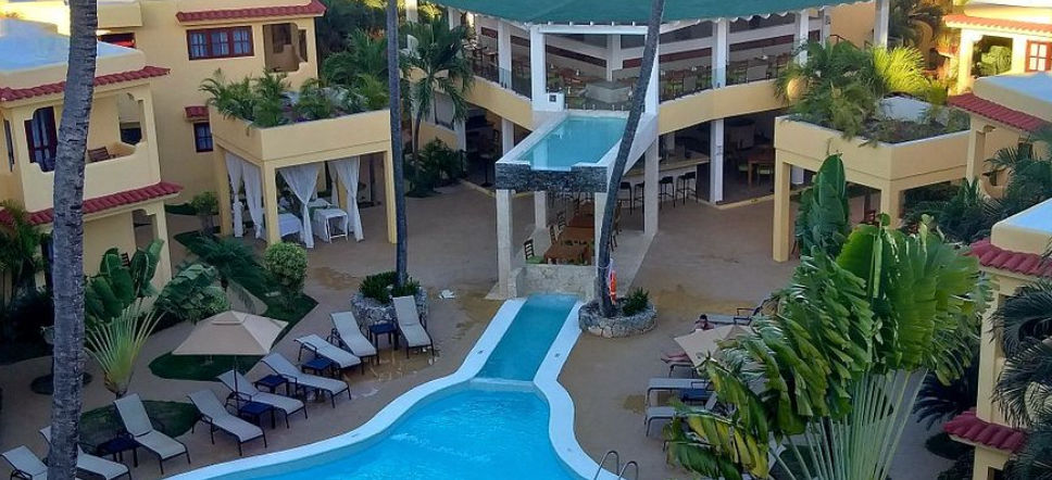 Aerial view of the central courtyard and pool area at Ducassi Suites Beach Club & Spa in Punta Cana, featuring lounge chairs, palm trees, and an elevated architectural glass-edge pool with a waterfall feature.