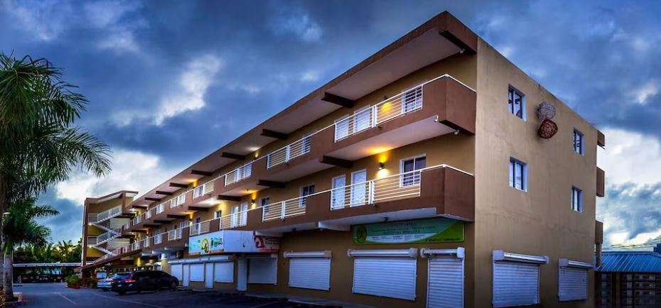 Exterior facade of My Home Hotel Punta Cana at dusk, a three-story brown building featuring white balconies, a parking area, and ground-floor commercial spaces.