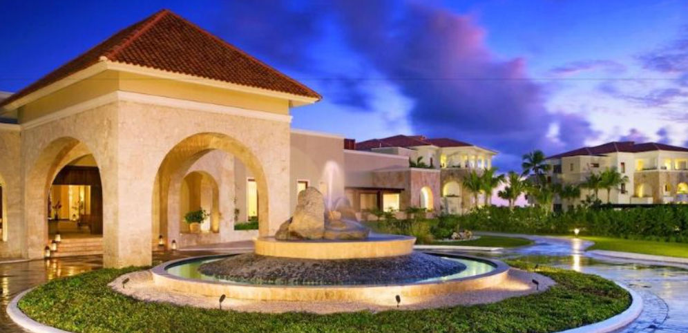 Illuminated main entrance of the luxury hotel Xeliter Golden Bear Lodge in Cap Cana, featuring a circular fountain, limestone arches, and residential villas at sunset.