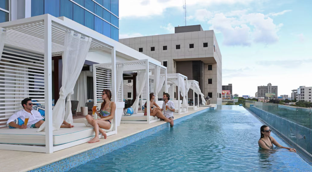 Night view of the Intercontinental Real Santo Domingo hotel showing its blue-lit infinity pool, outdoor terrace, and city traffic on Winston Churchill Avenue