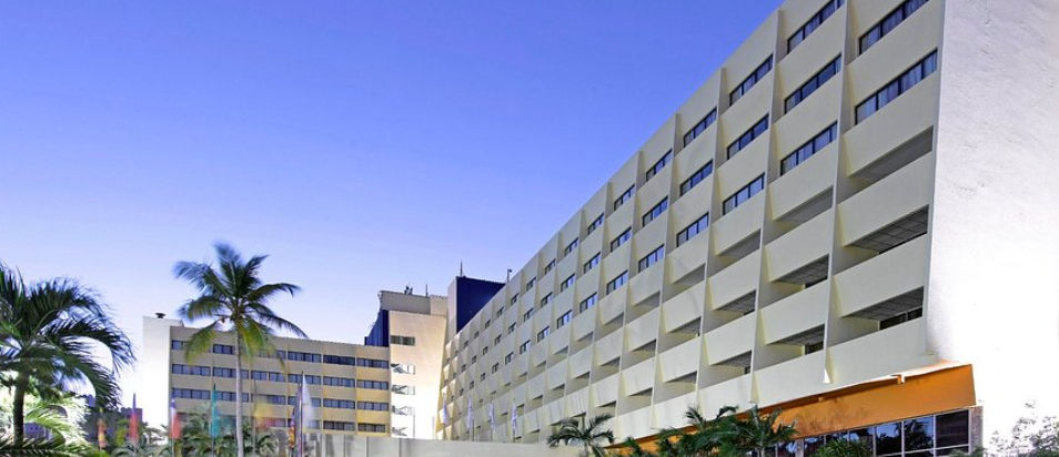 Exterior facade of the Dominican Fiesta Hotel & Casino in Santo Domingo under a clear blue sky with palm trees at the entrance.