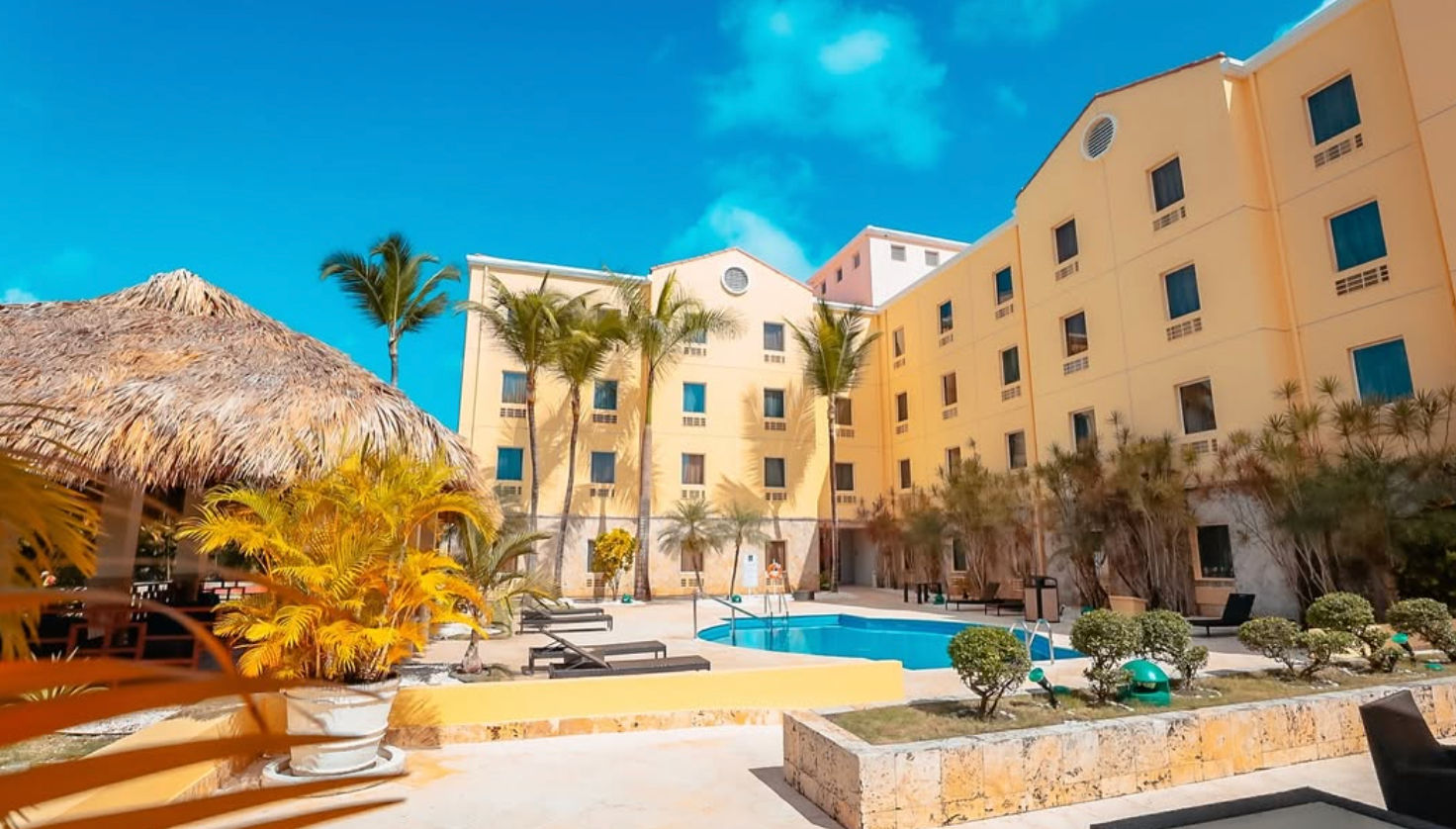 Exterior facade of the Quality Hotel Real Aeropuerto Santo Domingo featuring its light yellow building structure, red tile roofs, and the official Quality Hotel logo under a clear blue sky