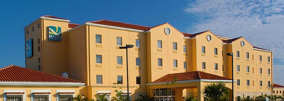 Exterior facade of the Quality Hotel Real Aeropuerto Santo Domingo featuring its light yellow building structure, red tile roofs, and the official Quality Hotel logo under a clear blue sky.