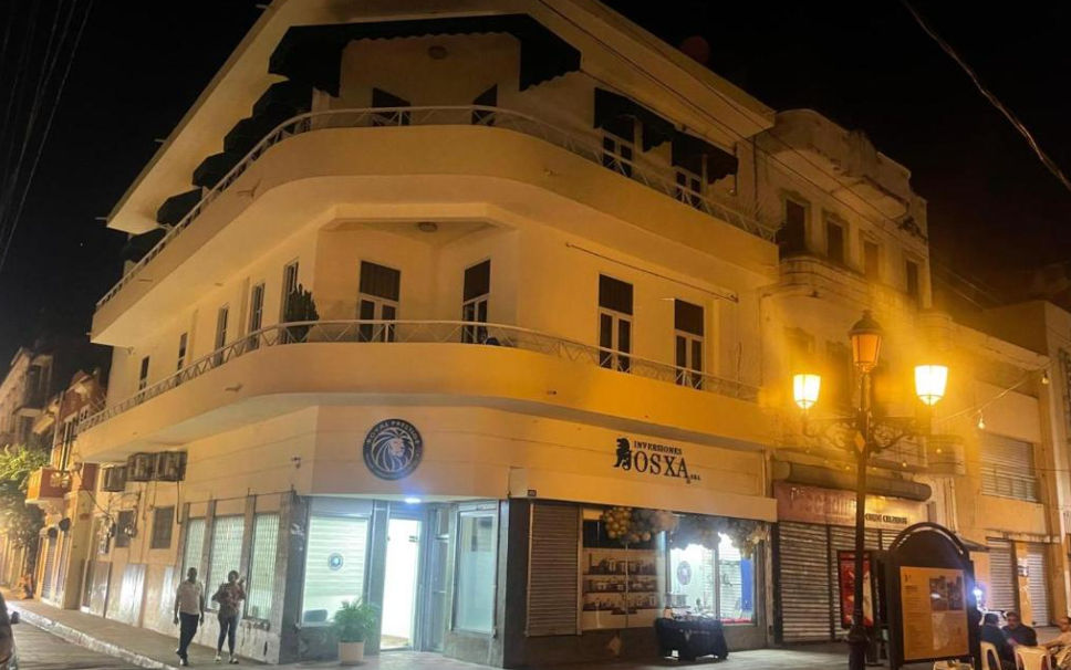 Nighttime view of a multi-story corner building with curved balconies and ground-floor commercial shops, illuminated by classic street lamps.