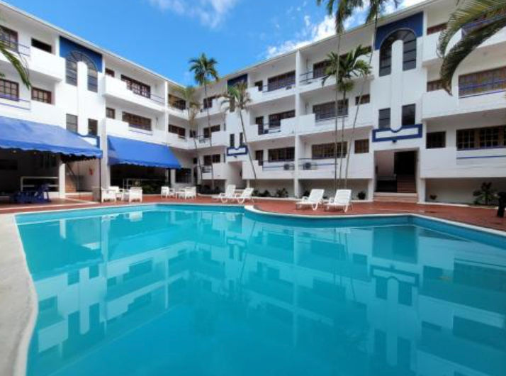 Vista de la piscina al aire libre y el patio central del Calypso Beach Hotel rodeado de palmeras, áreas de descanso y la fachada amarilla del edificio.