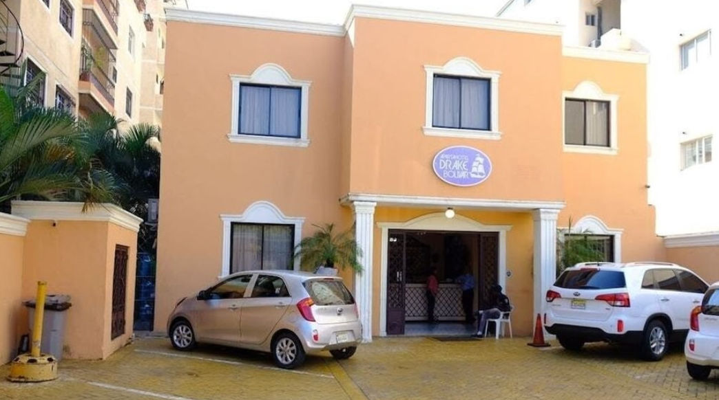 Main facade of Apartahotel Drake Boliva in Santo Domingo, showing the entrance of the two-story ochre building, the oval hotel sign, and vehicles parked in front.