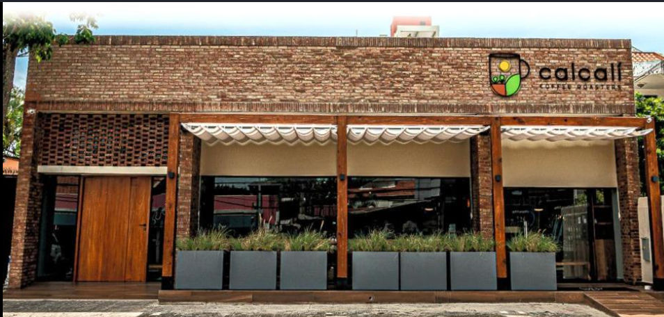 Fachada exterior de la cafetería Calcafí Coffee Roasters con pared de ladrillo, pilares de madera, toldos blancos y grandes ventanales de cristal detrás de modernas jardineras.