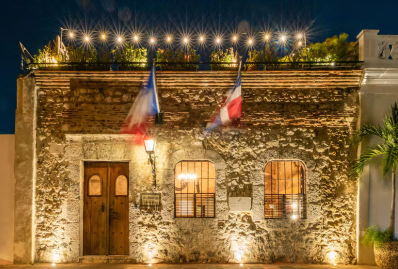Night view of the illuminated facade of El Beaterio Guest House in the Colonial City of Santo Domingo, featuring historic stone walls, an antique wooden door, and the flags of France and the Dominican Republic.